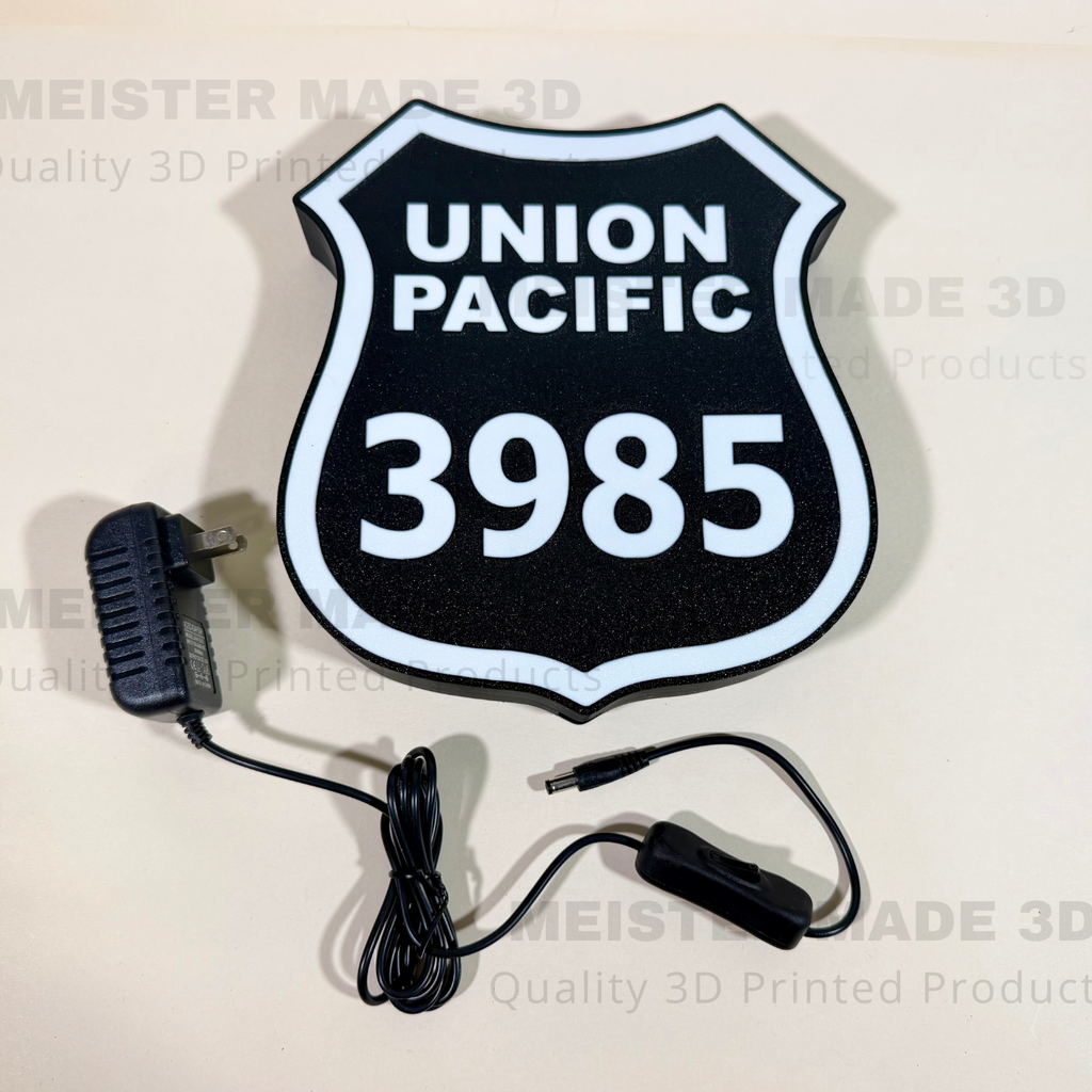 A 3D-printed Union Pacific 3985 railroad sign in the shape of a shield, shown with its power adapter with on/off switch on a light beige background.