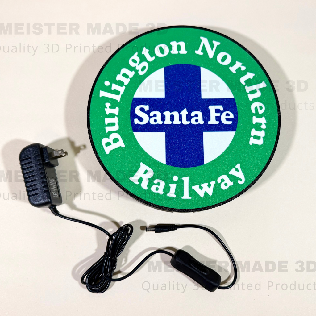 A circular green, white, and blue Burlington Northern Santa Fe Railway logo illuminated sign shown on a light background, with a black power adapter and cable placed below it.