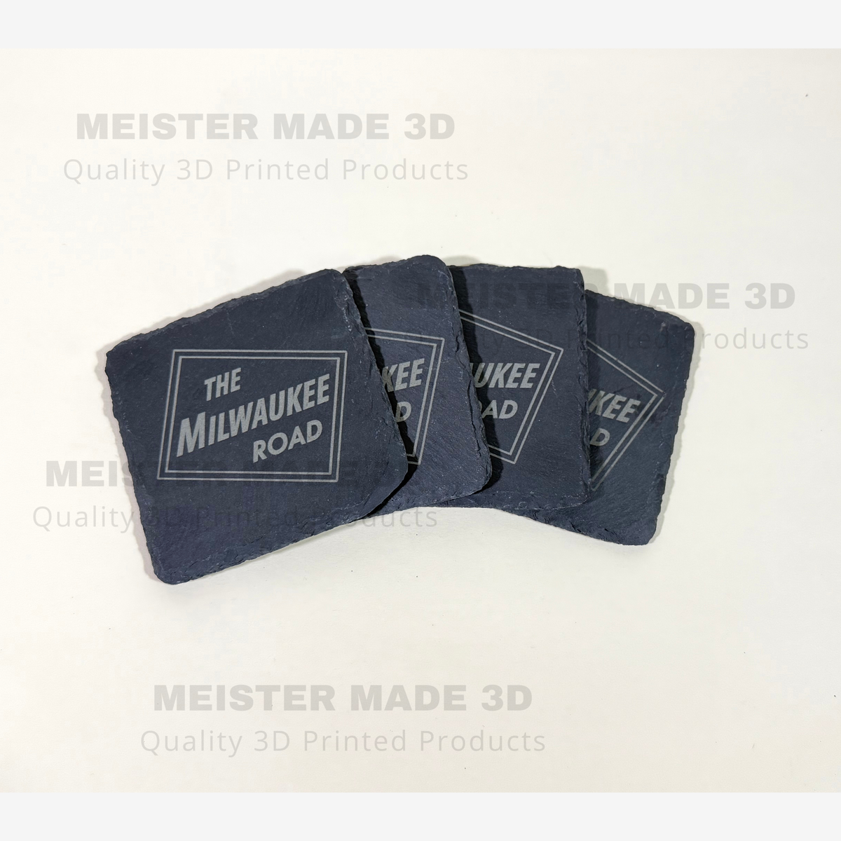 Four dark slate coasters with a white printed 'THE MILWAUKEE ROAD' logo, arranged in a fan shape on a light beige background with faint watermarks.