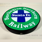 A circular 3D-printed sign featuring the Burlington Northern Santa Fe Railway logo, with a green outer ring, white lettering, a blue and white cross in the center, and a black raised edge, shown on a light beige background with faint watermarks.