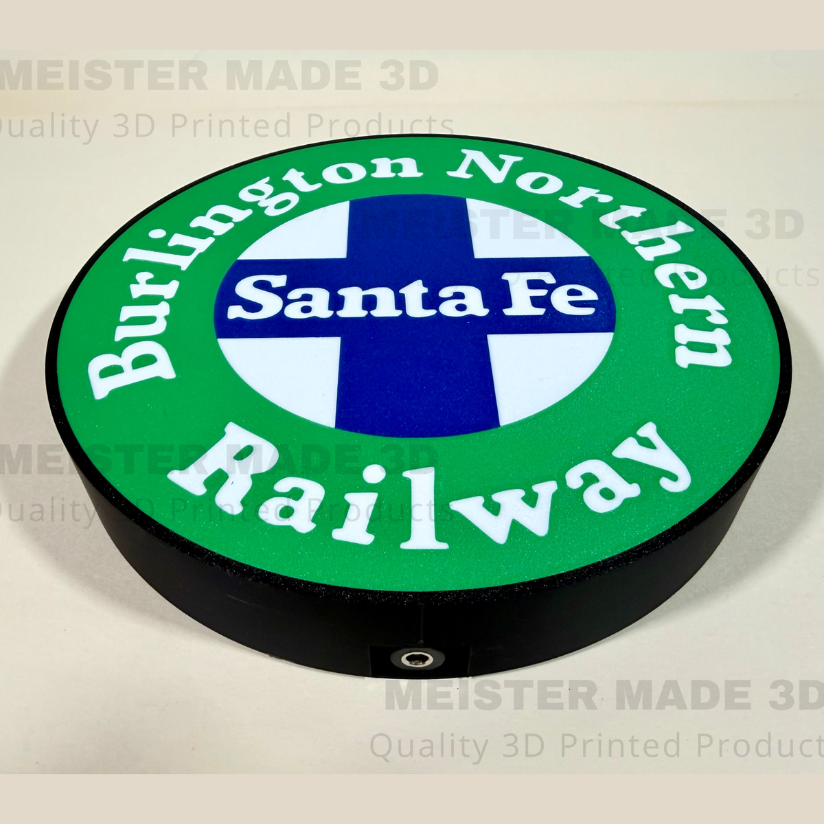 A circular 3D-printed sign featuring the Burlington Northern Santa Fe Railway logo, with a green outer ring, white lettering, a blue and white cross in the center, and a black raised edge, shown on a light beige background with faint watermarks.