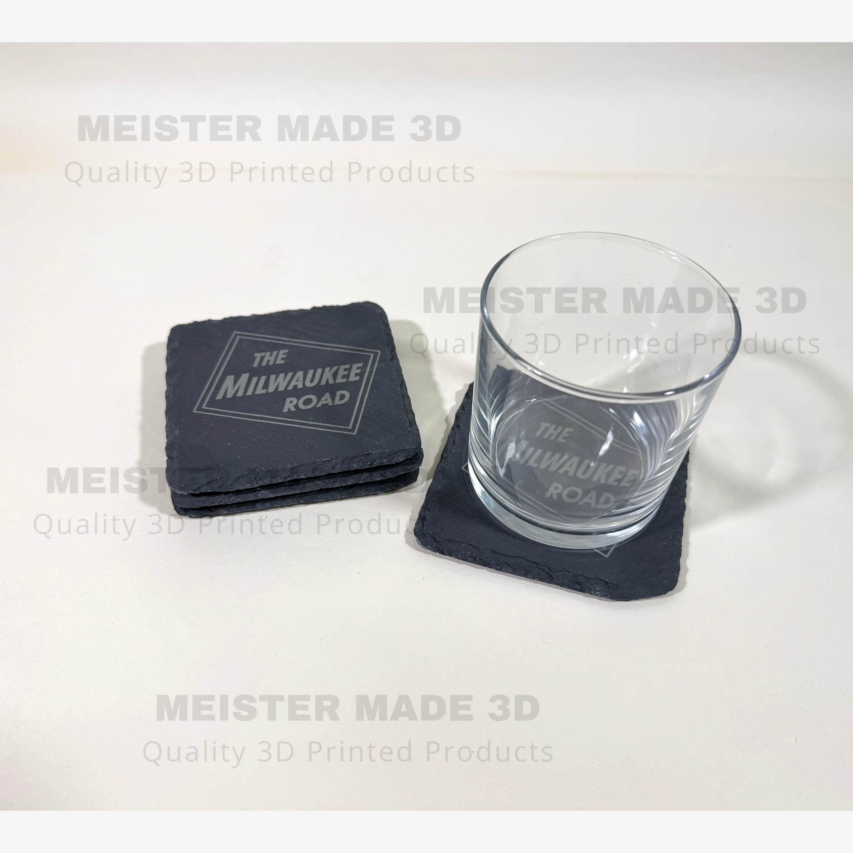 A clear glass and a stack of dark slate coasters printed with 'The Milwaukee Road' logo, shown on a light background with repeated 'MEISTER MADE 3D' watermark text.