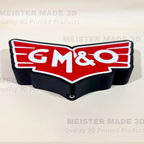 A 3D printed GM&O logo sign with a red and white top section and a black base, shown on a light beige background with 'MEISTER MADE 3D' watermarks.