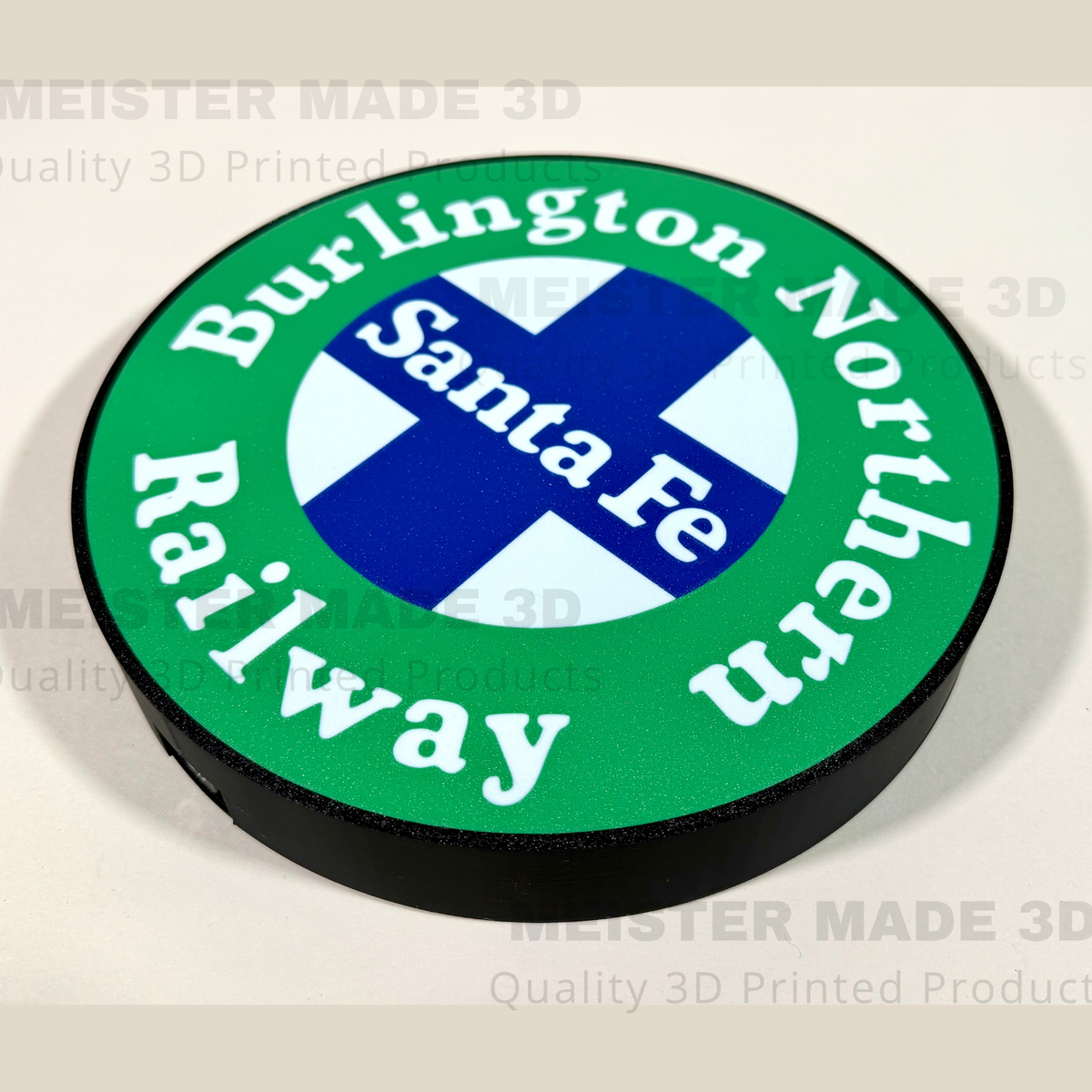A circular 3D-printed sign featuring the Burlington Northern Santa Fe Railway logo, with a green outer ring, white lettering, a blue and white cross in the center, and a black raised edge, shown on a light beige background with faint watermarks.
