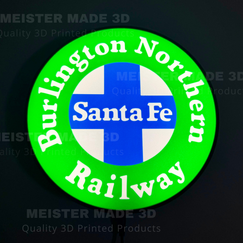 A circular 3D-printed illuminated sign for the Burlington Northern Santa Fe Railway, featuring a bright green outer ring with white lettering, a blue and white cross in the center, and the words 'Santa Fe' in white. The sign is photographed against a dark background with faint watermarks.