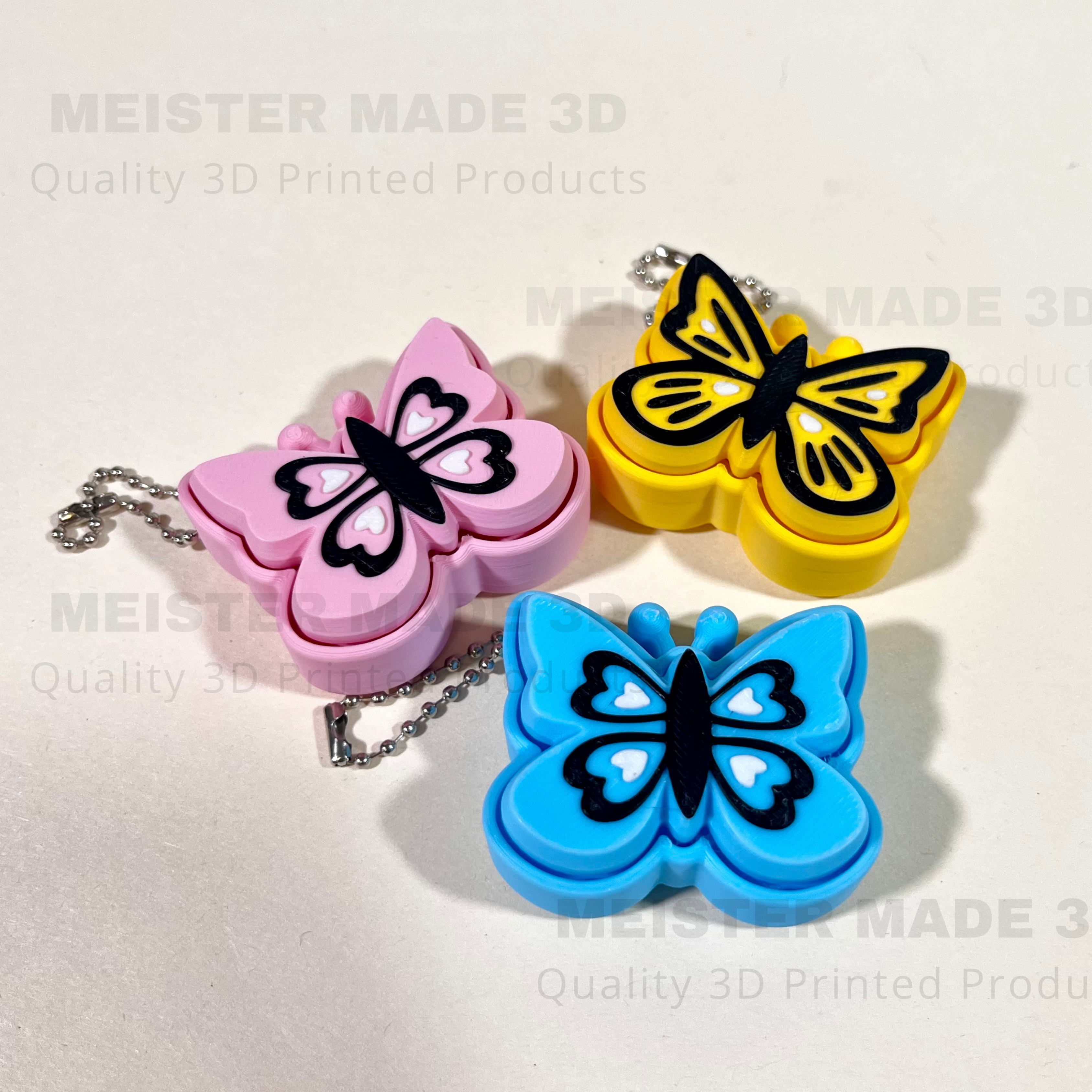 Three 3D-printed butterfly keychains in pink, yellow, and blue on a light gray background.