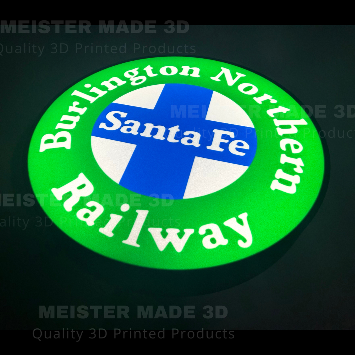 A circular 3D-printed illuminated sign for the Burlington Northern Santa Fe Railway, featuring a bright green outer ring with white lettering, a blue and white cross in the center, and the words 'Santa Fe' in white. The sign is photographed against a dark background with faint watermarks.